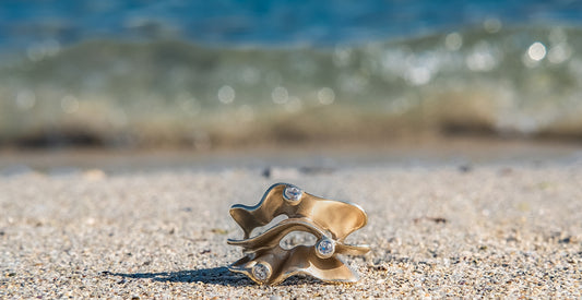 Anello Onde in bronzo oro sabbiato e zirconi di Roberta Risolo fotografato in riva al mare con i riflessi del sole e della sabbia dorata